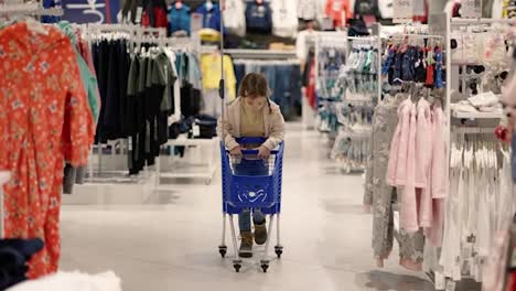 Little-girl-is-having-fun-in-a-clothes-store,-riding-a-shopping-cart