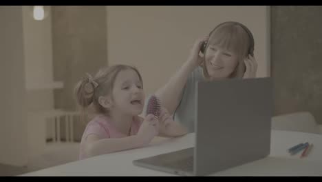 Kleines-Mädchen-Sitzt-Am-Schreibtisch-Am-Laptop-Und-Singt,-Während-Großmutter-In-Der-Nähe-Musik-über-Kopfhörer-Hört
