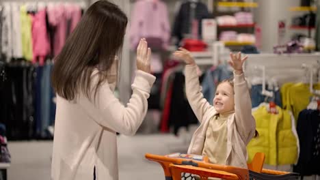 Well-done,-success-concept---girl-sits-in-a-supermarket-trolley-and-claps-her-hands-on-her-mother's-hands