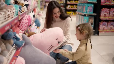 Mother-shows-big-soft-toy-to-her-daughter-in-children's-department-store,-both-smiling