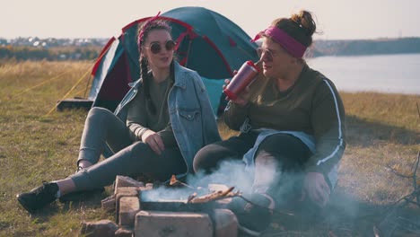 Glückliche-Mädchen-Trinken-Tee-Am-Brennenden-Lagerfeuer-In-Der-Nähe-Des-Blauen-Zeltes