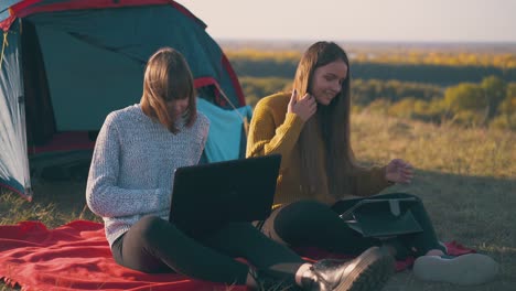 girls-use-tablet-and-laptop-sitting-at-tent-on-river-bank
