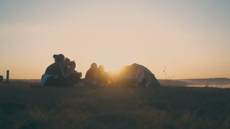 young-people-rest-at-burning-bonfire-on-river-bank-at-sunset