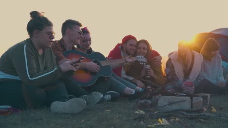young-people-sit-with-guitar-at-burning-bonfire-in-evening