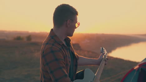 guy-plays-guitar-in-camp-against-river-and-setting-sun