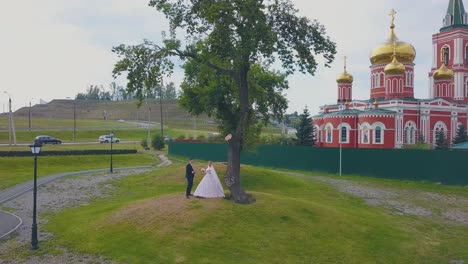 bride-and-groom-dance-near-tree-against-church-aerial-view