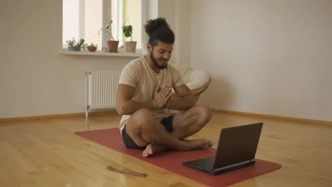 Bearded-man-talk-on-video-call-sit-on-yoga-mat-greets-in-namaste