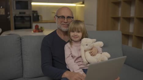 Grandfather-and-small-granddaughter-sitting-on-sofa-and-looking-to-the-camera