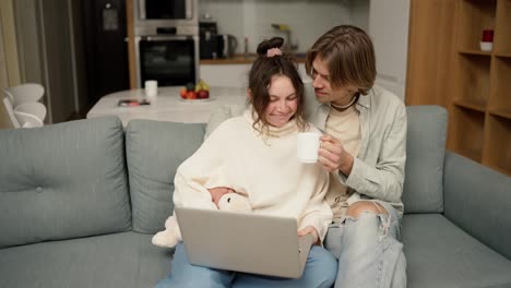 Young-adult-couple-talking-using-laptop-computer-at-home-sitting-on-couch,-watching-video