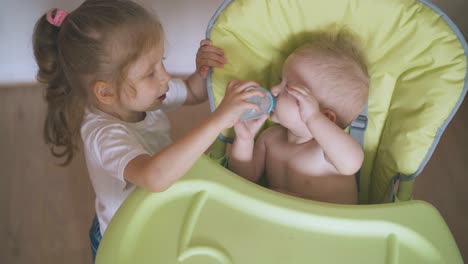 sister-strokes-baby-giving-water-to-brother-in-highchair