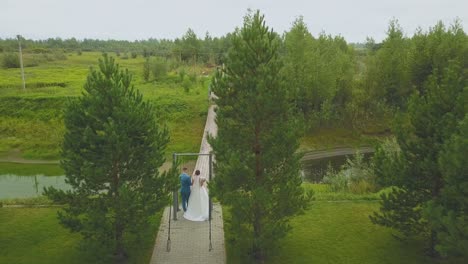 newlywed-couple-walks-to-wooden-bridge-aerial-backside-view