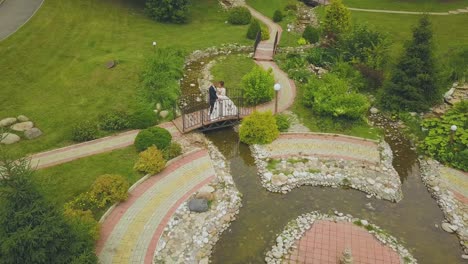 Frischvermähltes-Paar-Auf-Brücke-über-Bach-Im-Park,-Luftaufnahme