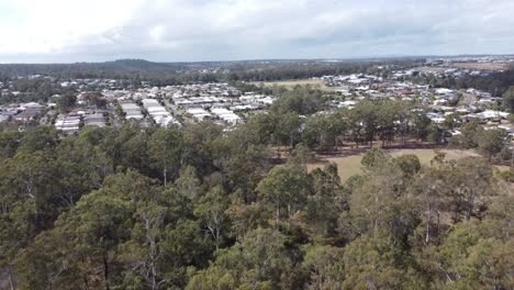 Drone-flying-over-bushland-towards-a-green-field-and-residential-estate-in-the-background