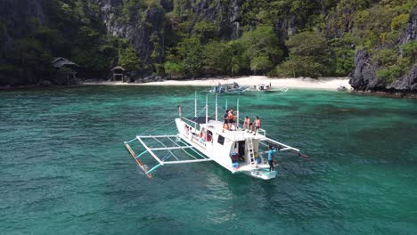Island-Hopping-tourists-on-top-deck-of-traditional-outrigger-boat-fronting-tropical-Serenity-Beach-on-Cadlao-Island,-El-Nido-Philippines