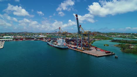 Shipping-container-port-with-a-small-boat-docked-and-2-cranes,-cinematic-aerial-drone-tilt-down