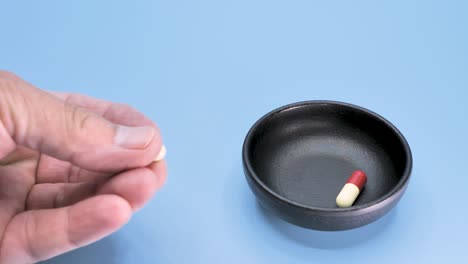 A-man's-hand-takes-one-pill-from-two,-from-a-black-bowl-standing-on-a-blue-surface