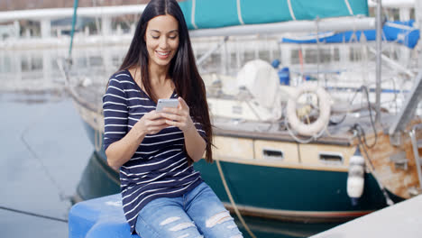 Young-woman-sitting-on-a-quay-checking-her-mobile
