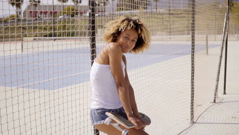 Young-woman-with-skateboard-leans-against-fence