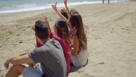Backs-of-teens-sitting-on-sandy-beach-outdoors