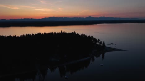 Drone-shot-of-Herron-Island's-silhouette-at-sunset-with-a-glowing-set-of-mountains-off-in-the-distance