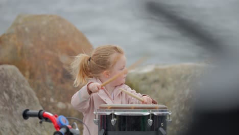 Young-ginger-hair-toddler-girl-bangs-on-drums-by-the-beach,-during-the-70th-anniversary-of-currach-racing-in-Salthill,-Galway-Ireland