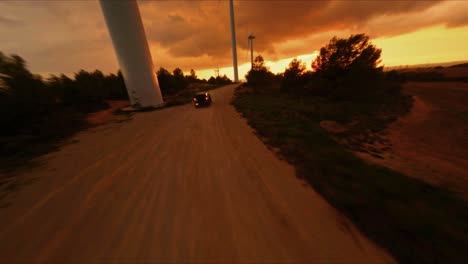 FPV-aerial-following-a-black-Hummer-driving-through-a-wind-turbine-farm-at-sunset