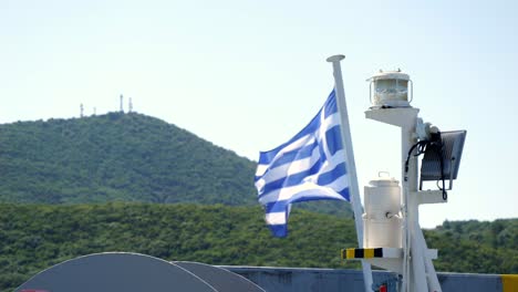 Bandera-Griega-Izada-En-Un-Mástil,-Con-Las-Montañas-De-La-Ciudad-De-Igoumenitsa-Al-Fondo