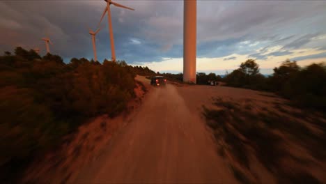 FPV-aerial-drone-following-a-black-Hummer-driving-under-wind-turbines-along-a-dirt-road-at-sunset