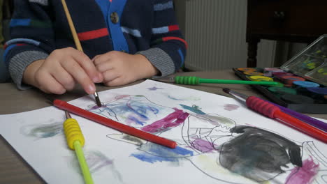 A-medium-close-up-of-a-young-boy-sat-a-desk-using-a-watercolour-paint-brush