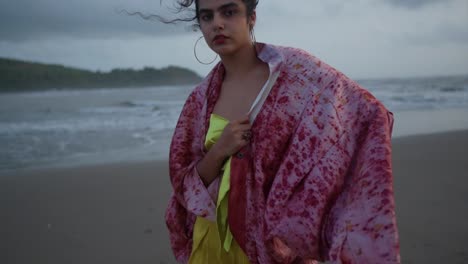 A-woman-with-dark-hair-in-a-red-cardigan-and-golden-dress-standing-on-the-beach-staring-at-the-camera