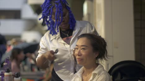 Asian-woman-getting-fresh-hairdo-by-African-woman-hair-stylist-at-indoor-ethnic-cultural-festival,-filmed-as-slow-motion-medium-close-up-shot-in-handheld-style