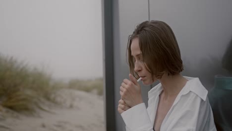 cinematic-video-of-a-young-and-stressed-pretty-woman-smoking-a-cigarette-outside-near-the-white-sand-beach-behind-a-black-building