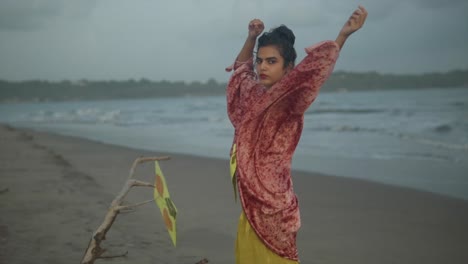 Slow-motion-shot-of-a-woman-in-a-golden-dress-and-red-cardigan-standing-on-the-beach-with-her-hands-in-the-air
