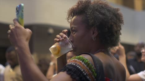 African-woman-drinking-alcoholic-beverage-while-using-cellphone-to-film-and-live-stream-at-outdoor-ethnic-cultural-event,-filmed-as-close-up-in-slow-motion