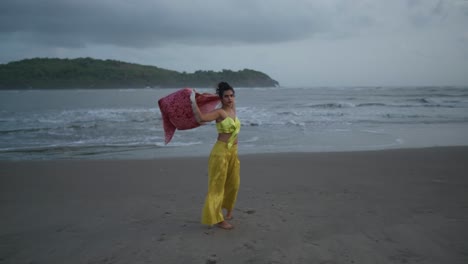 Cinematic-footage-of-Indian-fashion-model-wearing-a-yellow-beautiful-outfit-and-a-red-silk-jacket-on-the-beach-in-the-Wind-in-Slow-Motion-in-Goa-India