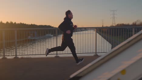 Side-tracking-shot-of-man-a-running-on-a-bridge-during-golden-hour-with-parallaxing-metal-bridge-beams