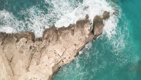 Aerial-overhead-drone-footage-unveils-hidden-turquoise-bay,-where-white-waves-crash-on-the-beach-and-a-vibrant-brown-rock-off-Gozo's-coast,-sea-spray-soaring