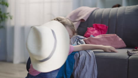Little-girl-plays-with-hat-putting-into-suitcase-on-sofa