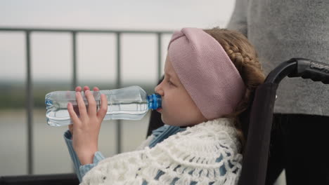 Thirsty-girl-drinks-water-sitting-in-wheelchair-near-mom