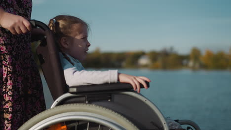 Preschooler-with-braids-sits-in-wheelchair-on-coast-of-lake