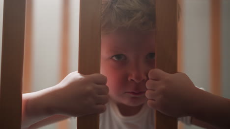Scared-little-boy-grabs-wooden-bars-of-crib-with-hands