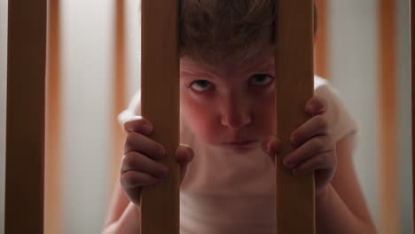 Offended-little-boy-looks-through-wooden-grid-of-baby-cradle