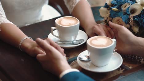 Newlyweds-hold-hands-while-sitting-in-a-cafe-sitting-at-a-table-on-which-there-are-two-mugs-of-coffee-close-up