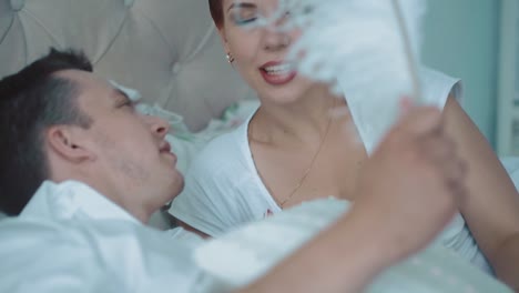 Young-beautiful-couple-having-fun-in-bed-they-are-fighting-with-pillows-he-tickles-her-with-a-big-feather-close-up