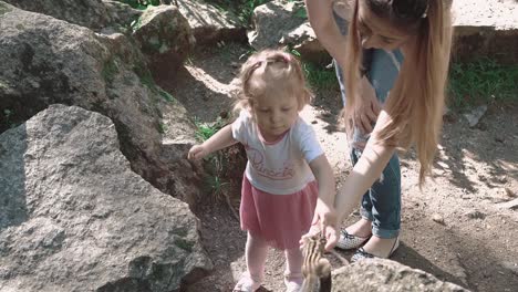 Young-beautiful-mother-with-her-daughter-feeding-a-chipmunk-in-the-woods-among-stones-3