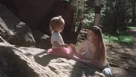 Young-beautiful-mother-and-daughter-talking-in-a-forest-near-big-stone