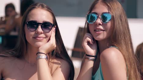 Two-young-attractive-hot-girls-wearing-sunglasses-are-sitting-at-table-on-beach