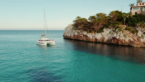 Catamarán-Navegando-En-El-Mar-Azul-Con-Casas-En-El-Acantilado-En-Cala-Llombards,-Mallorca,-España