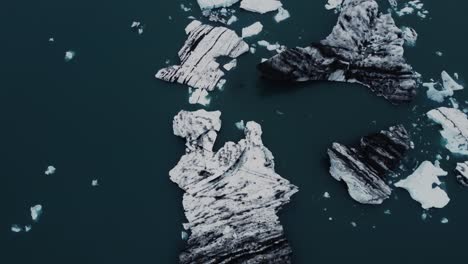 Drohnenaufnahme-Wunderschöner-Schwarz-weißer-Eisberge,-Die-In-Einer-Lagune-In-Island-Schwimmen