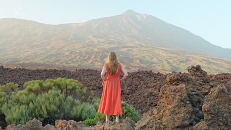 Girl-in-orange-summer-dress-standing-on-lava-rocks,-looking-at-volcano-Teide